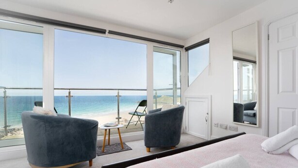 A bedroom with a large window overlooking the beach, two armchairs, a small table, and a bed with a pink and white striped bedspread. - Trewirgie - Central with sea views (St Ives)