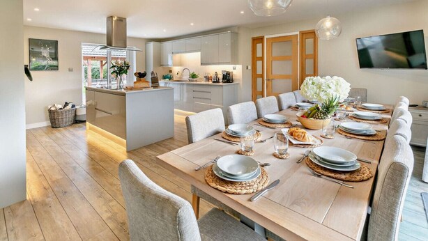 A modern kitchen with a central island, a dining area with a long table set for six, and a TV mounted on the wall.
