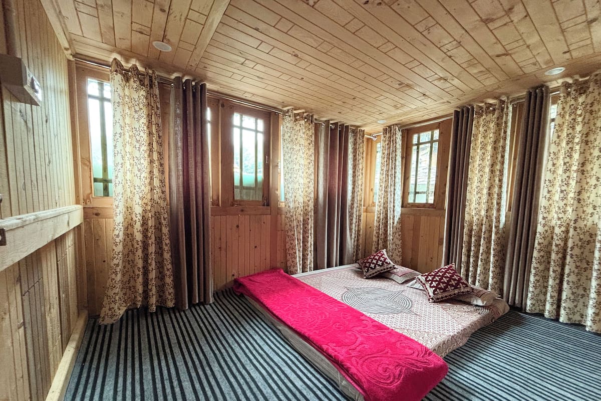 A room with wooden walls and ceiling, a bed with a red blanket, patterned pillows, and striped carpeting.