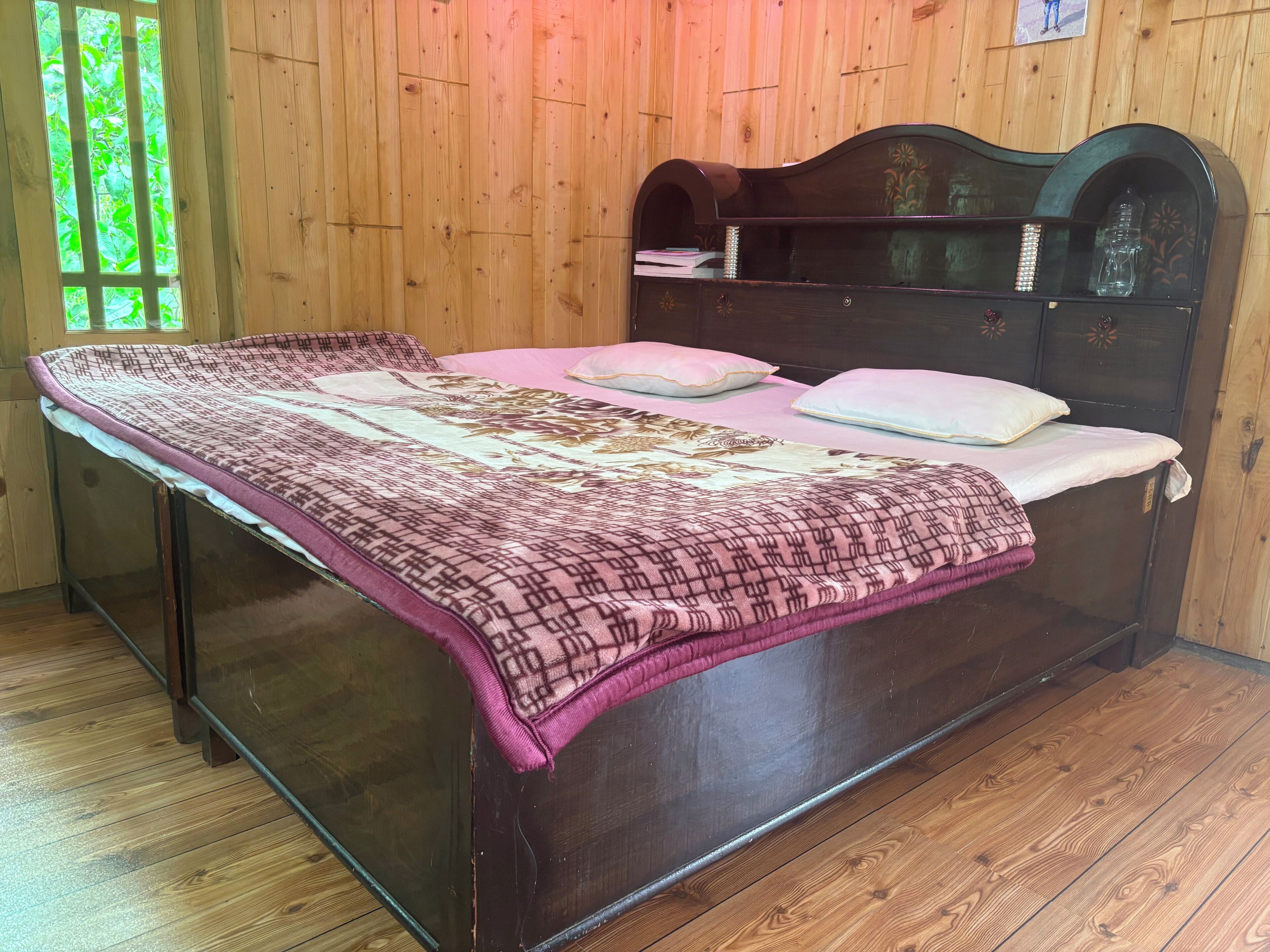A bed with a patterned bedspread, a wooden headboard, and a window with green foliage outside.