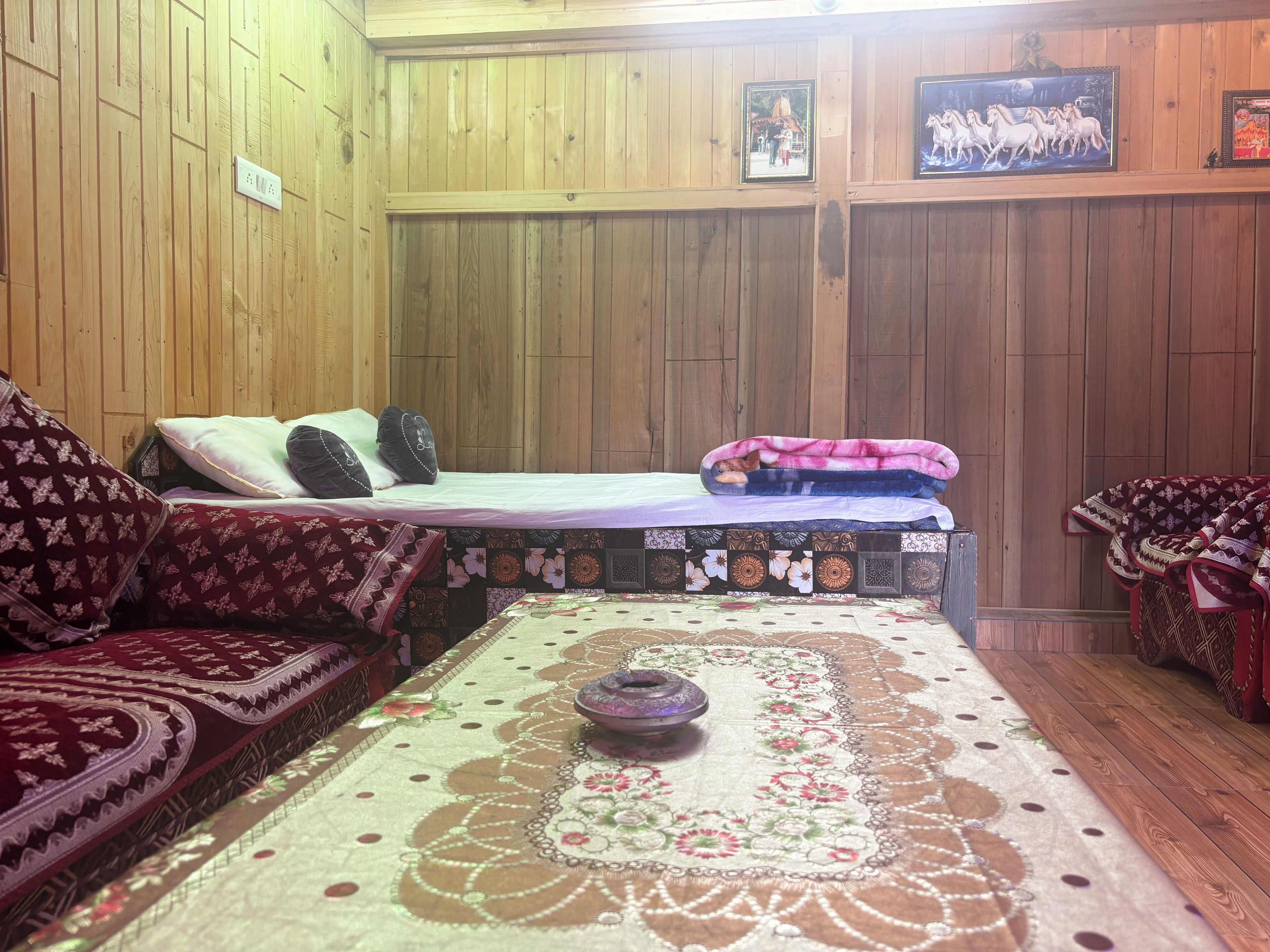 A room with wooden walls, a bed with a patterned bedspread, a sofa with a floral cover, and a decorative tablecloth.