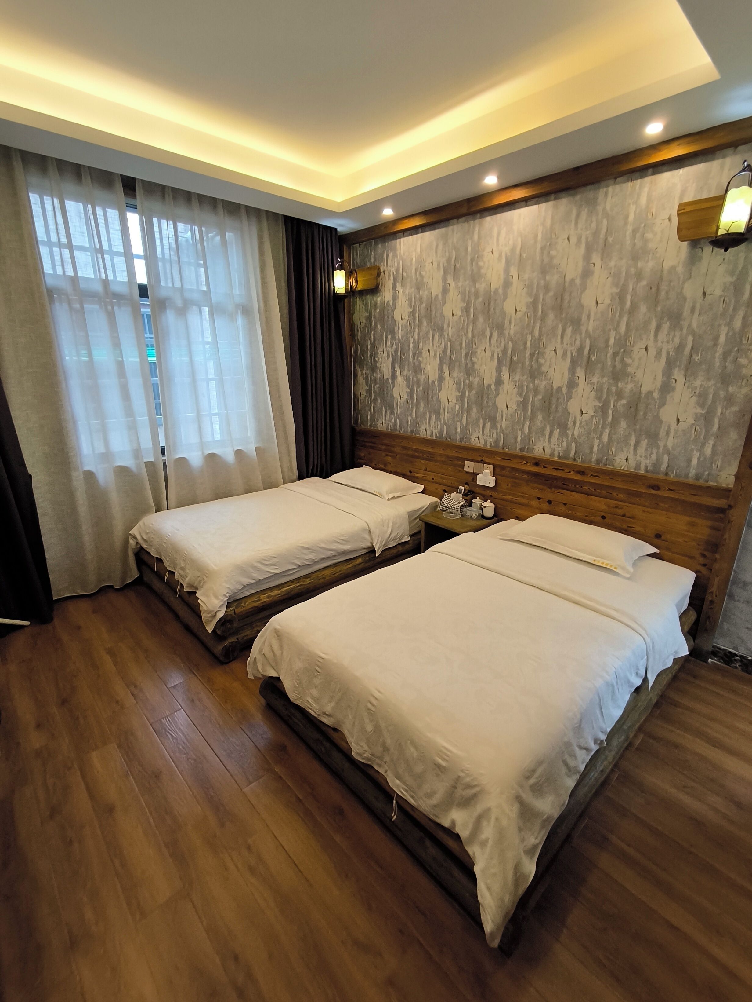 Quarto de hotel com duas camas individuais, piso de madeira, uma janela grande com cortinas leves e parede texturizada.