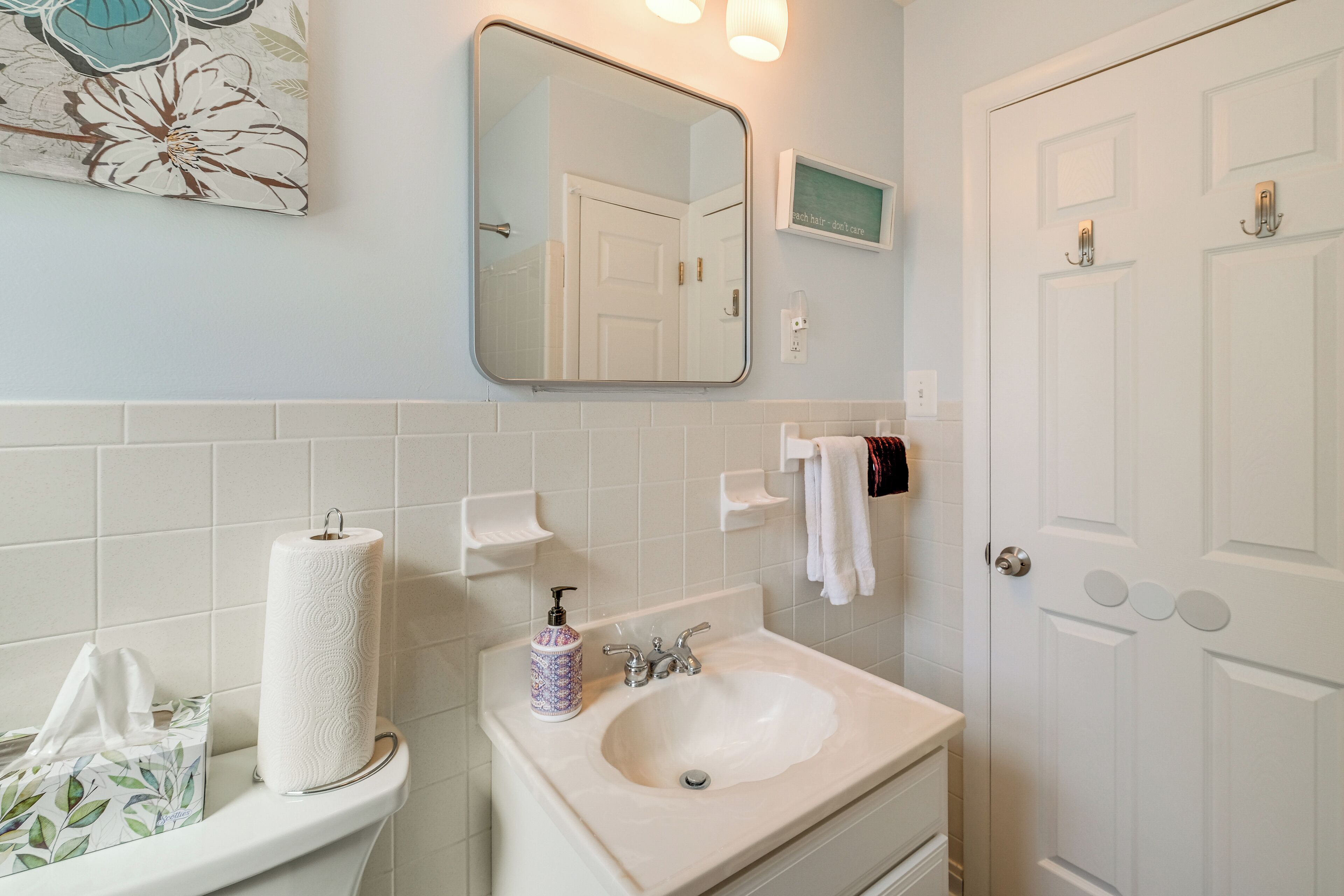 A bathroom with a toilet, sink, and mirror. There is a towel rack and a framed picture on the wall.