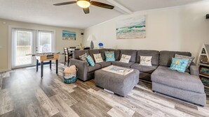 Un salon avec un canapé d’angle gris, un pouf et une table de baby-foot. On y trouve également un ventilateur de plafond, un tableau représentant une scène de plage et une fenêtre avec des stores.