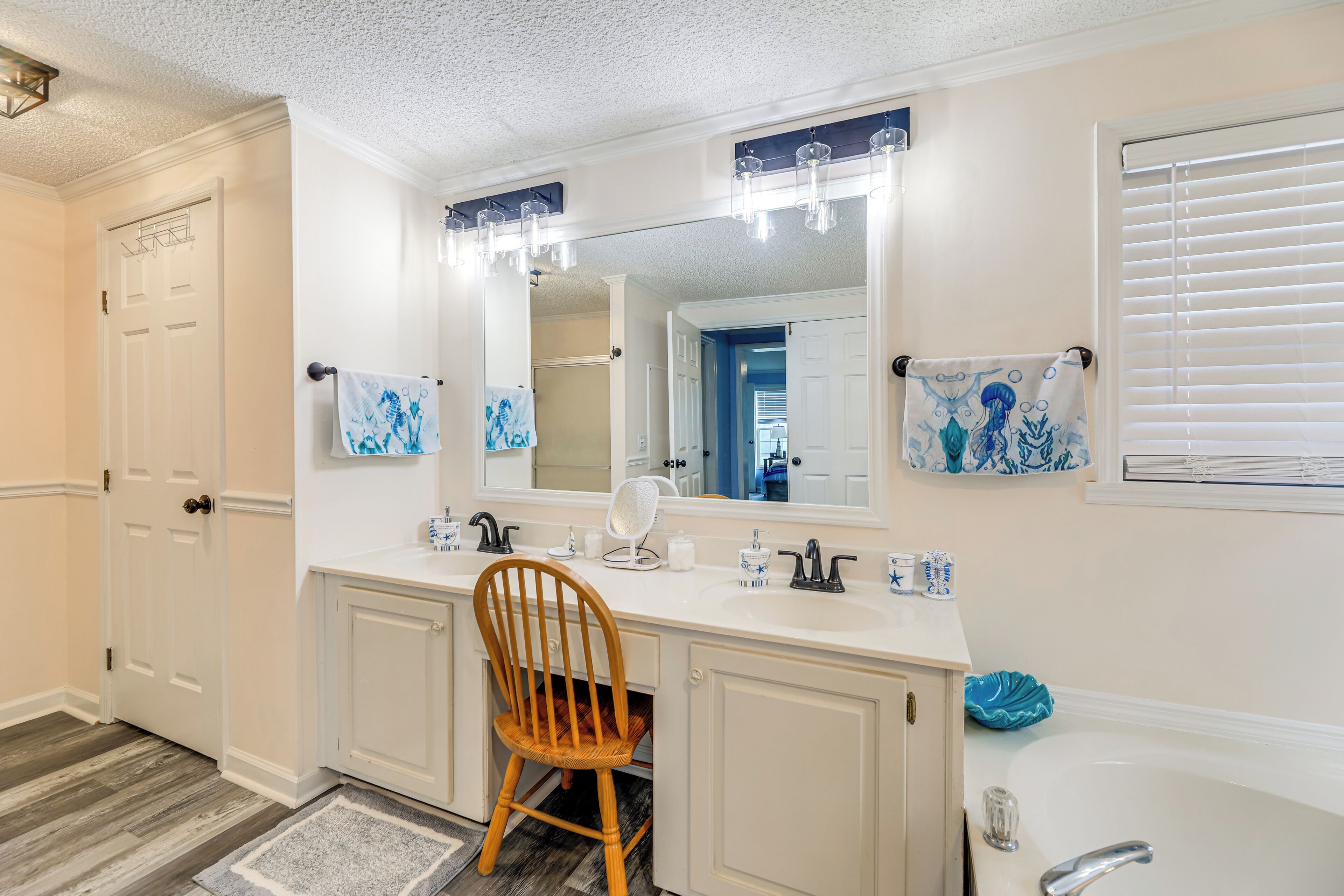 A bathroom with a white vanity, a large mirror, a wooden chair, a bathtub, and a window with blinds.