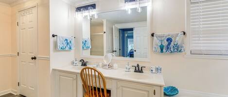 A bathroom with a white vanity, a large mirror, a wooden chair, a bathtub, and a window with blinds.