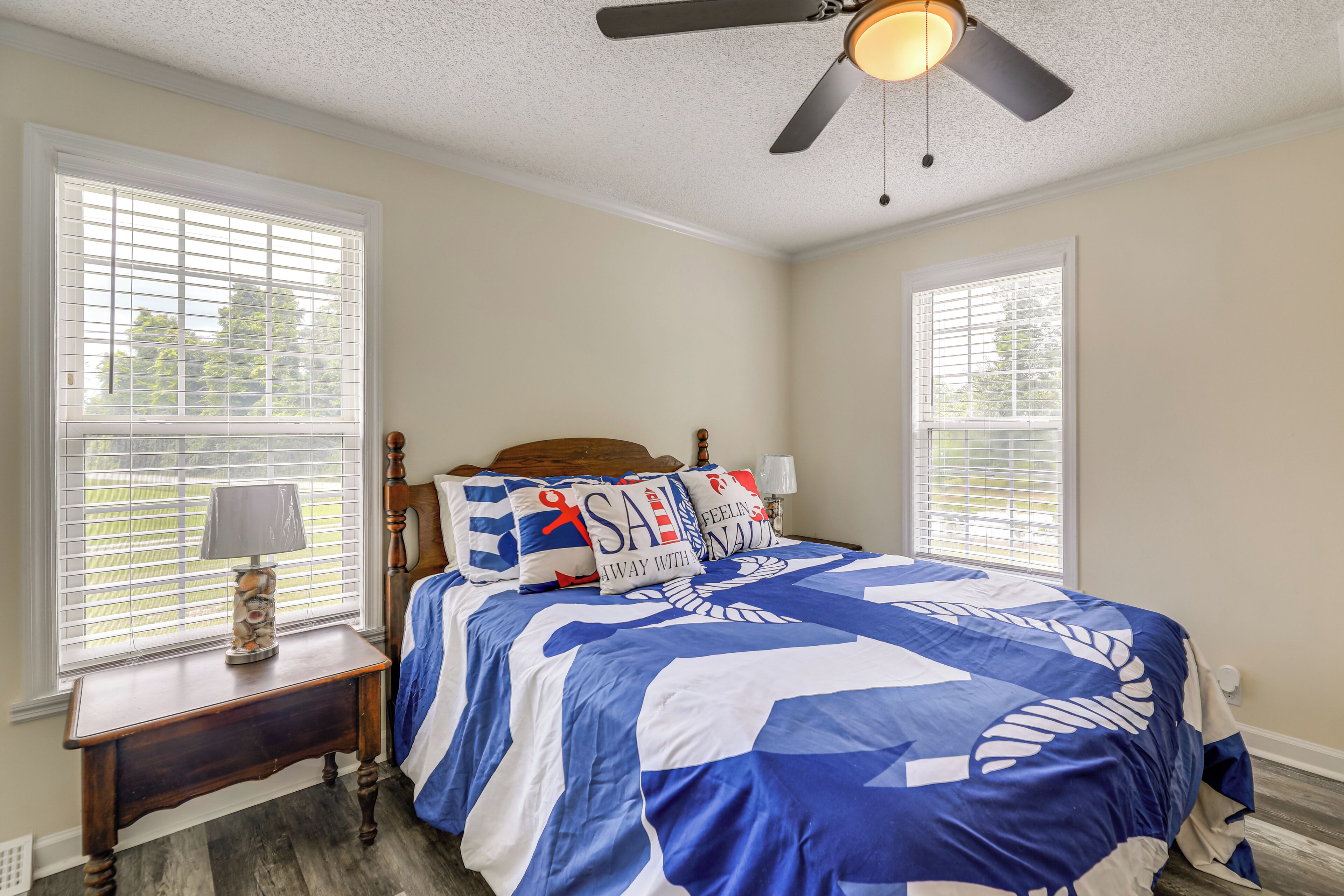 A bedroom with a bed, two windows, a nightstand, a lamp, and a ceiling fan.