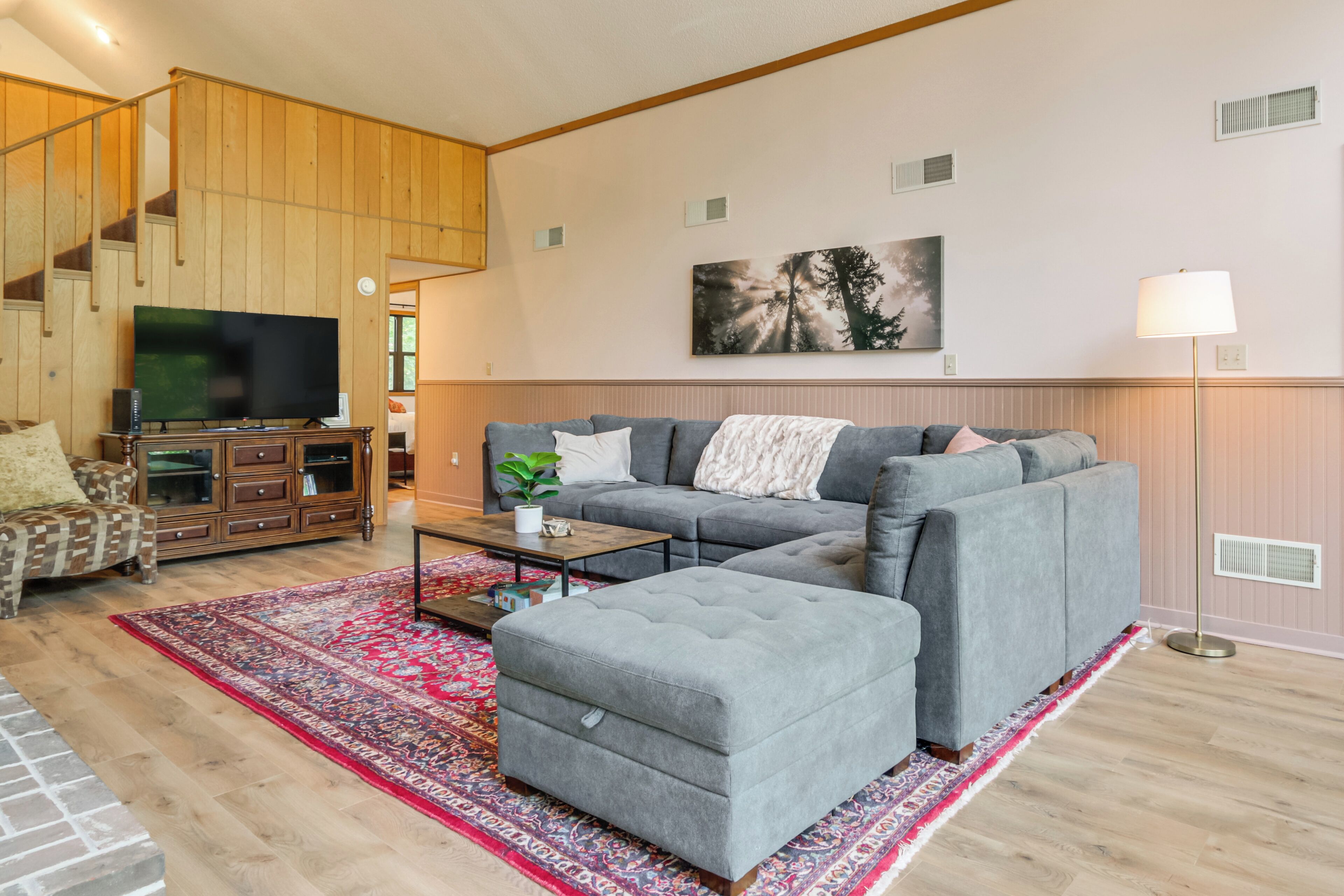 Una sala de estar moderna con un sofá seccional gris, un televisor de pantalla plana, un mueble de madera y un tapete rojo estampado.