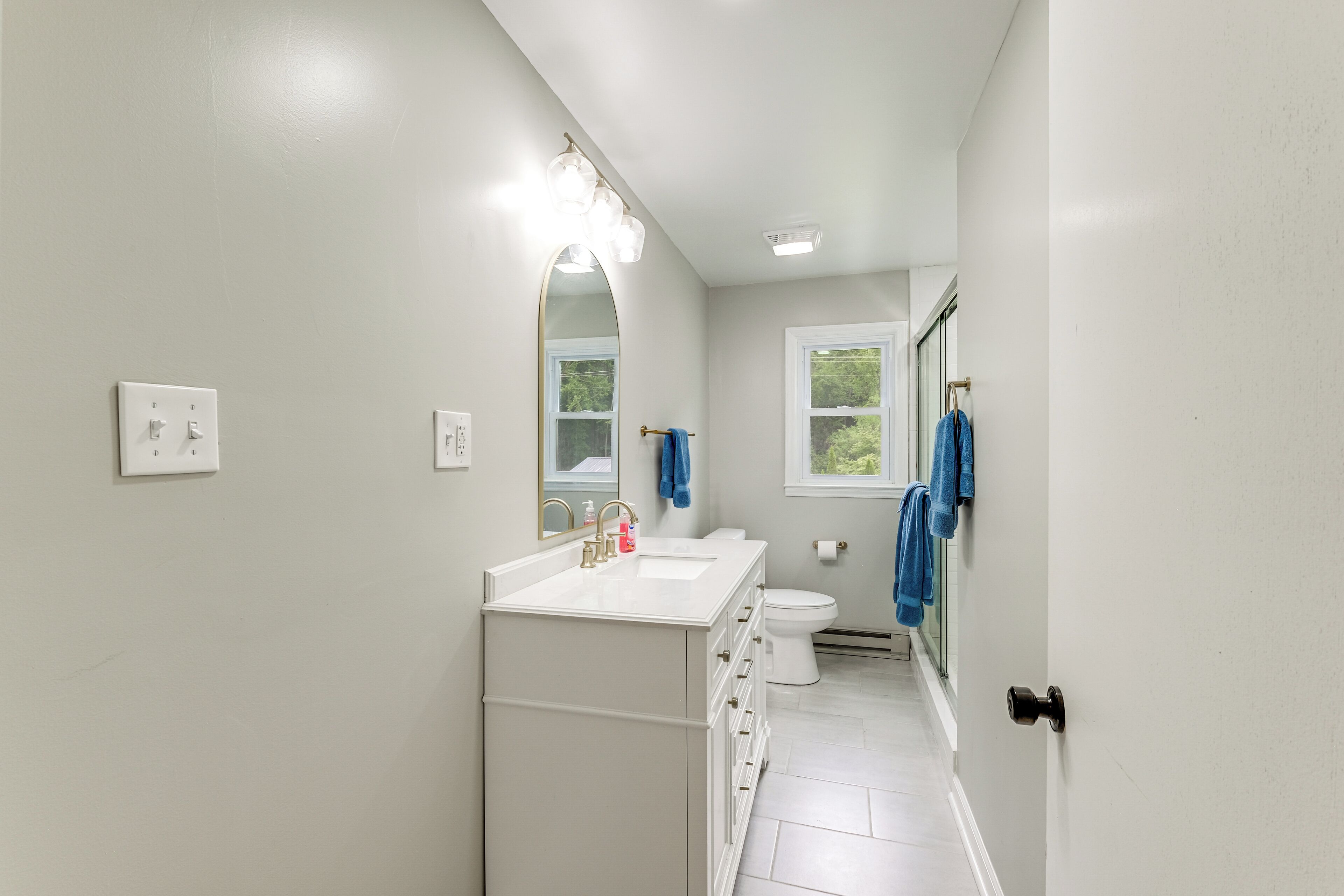 Un baño estrecho con un lavabo blanco, un espejo y un inodoro. Hay toallas colgadas en la pared y una ventana con vistas a árboles.