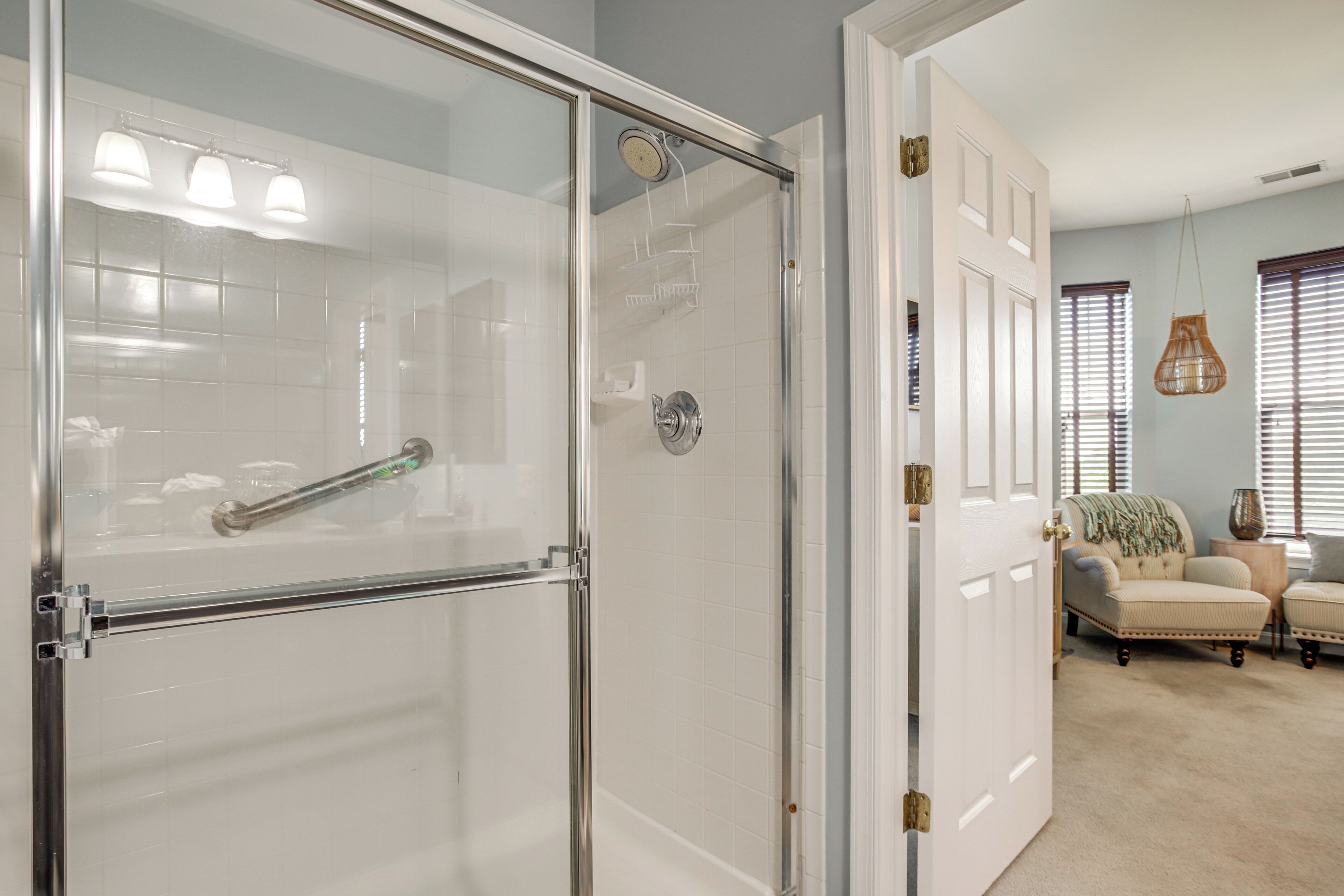A bathroom with a glass shower enclosure, a white tiled wall, and a door leading to a living area with a beige carpet, a chair, and a window with blinds.