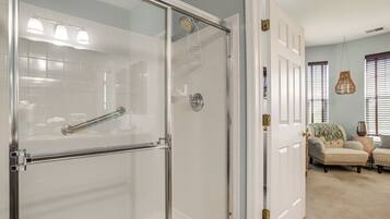 A bathroom with a glass shower enclosure, a white tiled wall, and a door leading to a living area with a beige carpet, a chair, and a window with blinds.
