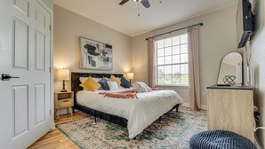A bedroom with a bed, bedside lamps, a ceiling fan, a window with curtains, a wooden dresser, and a rug.