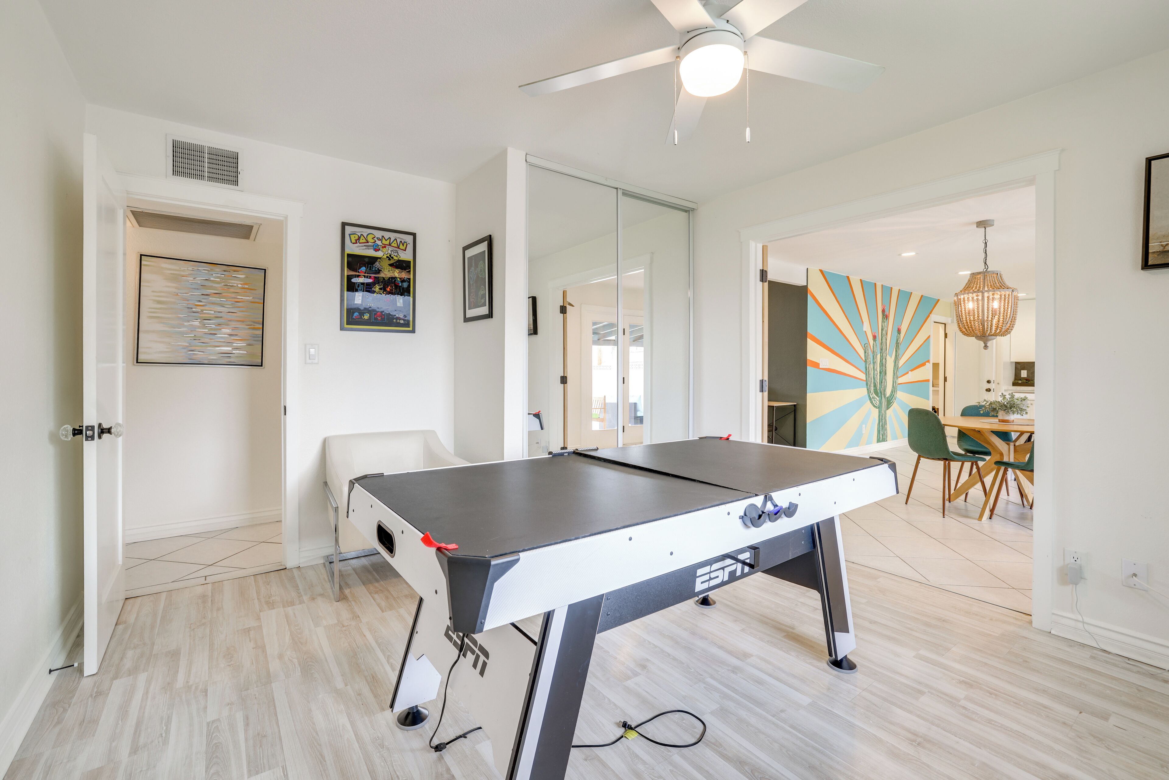 Quarto moderno com mesa de pebolim, ventilador de teto e uma área de jantar visível por uma porta aberta.