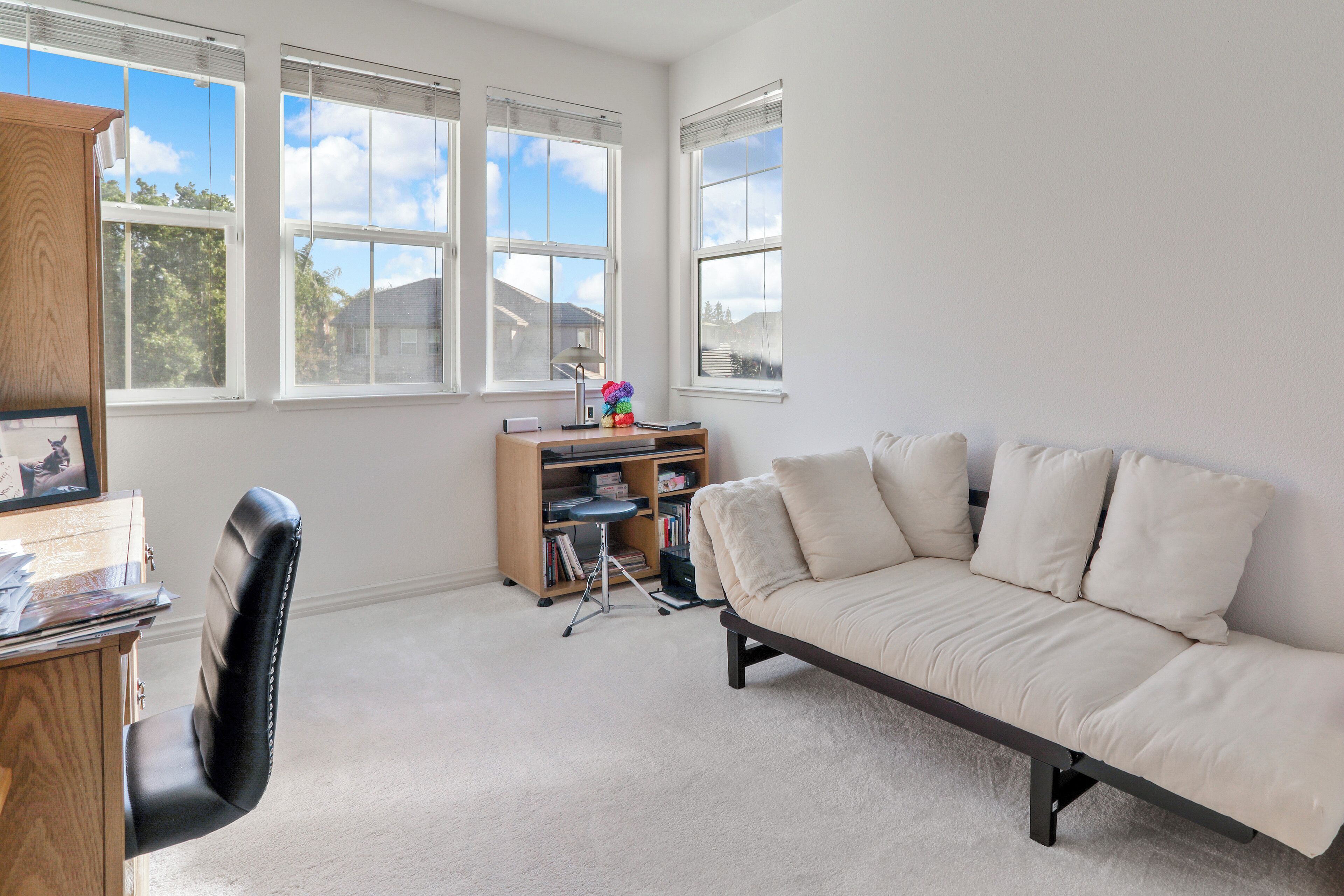 A living room with a beige sofa, a wooden cabinet, a desk with a chair, and a bookshelf.