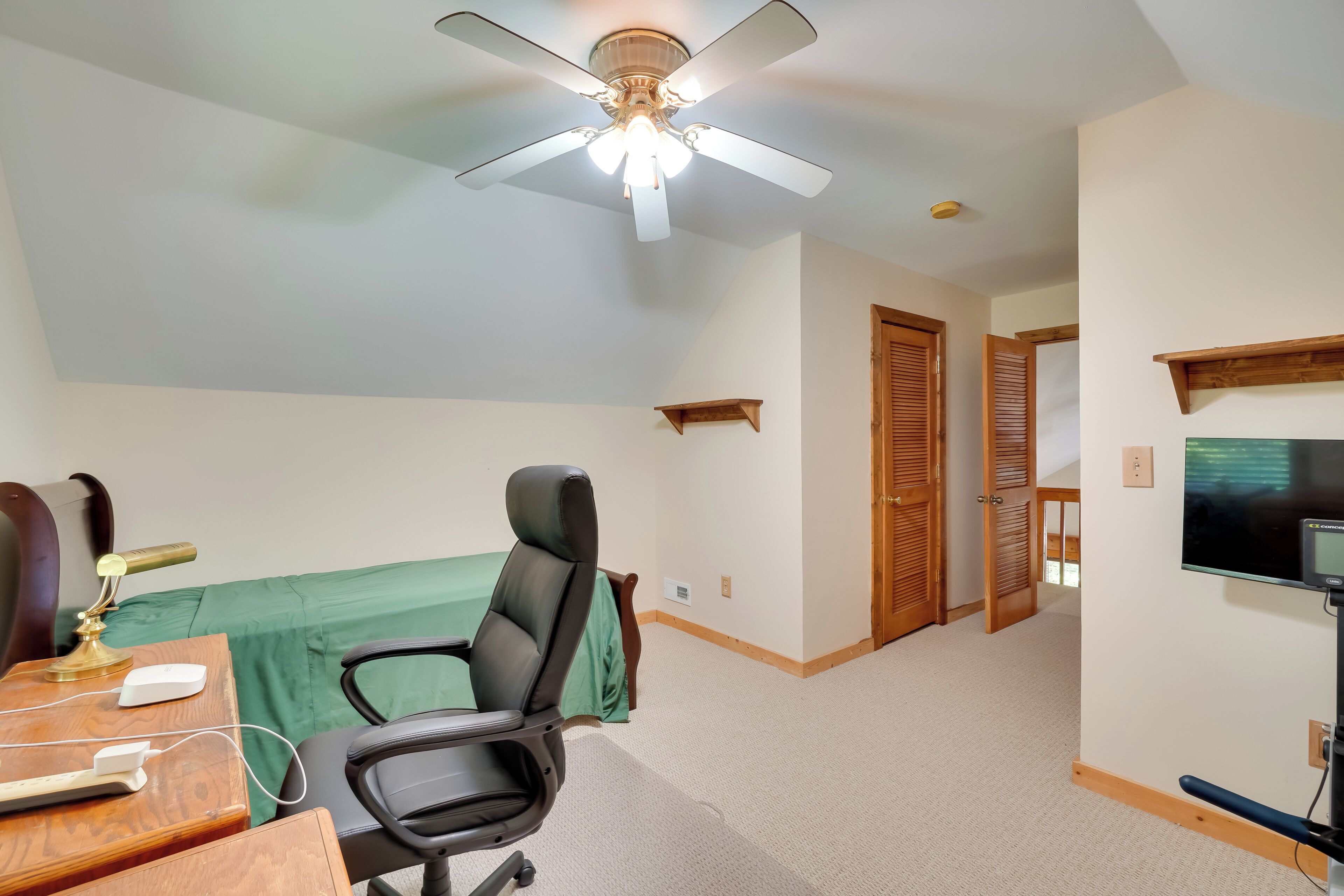A room with a ceiling fan, a desk with a lamp, a chair, a bed with a green cover, a television, and wooden doors.