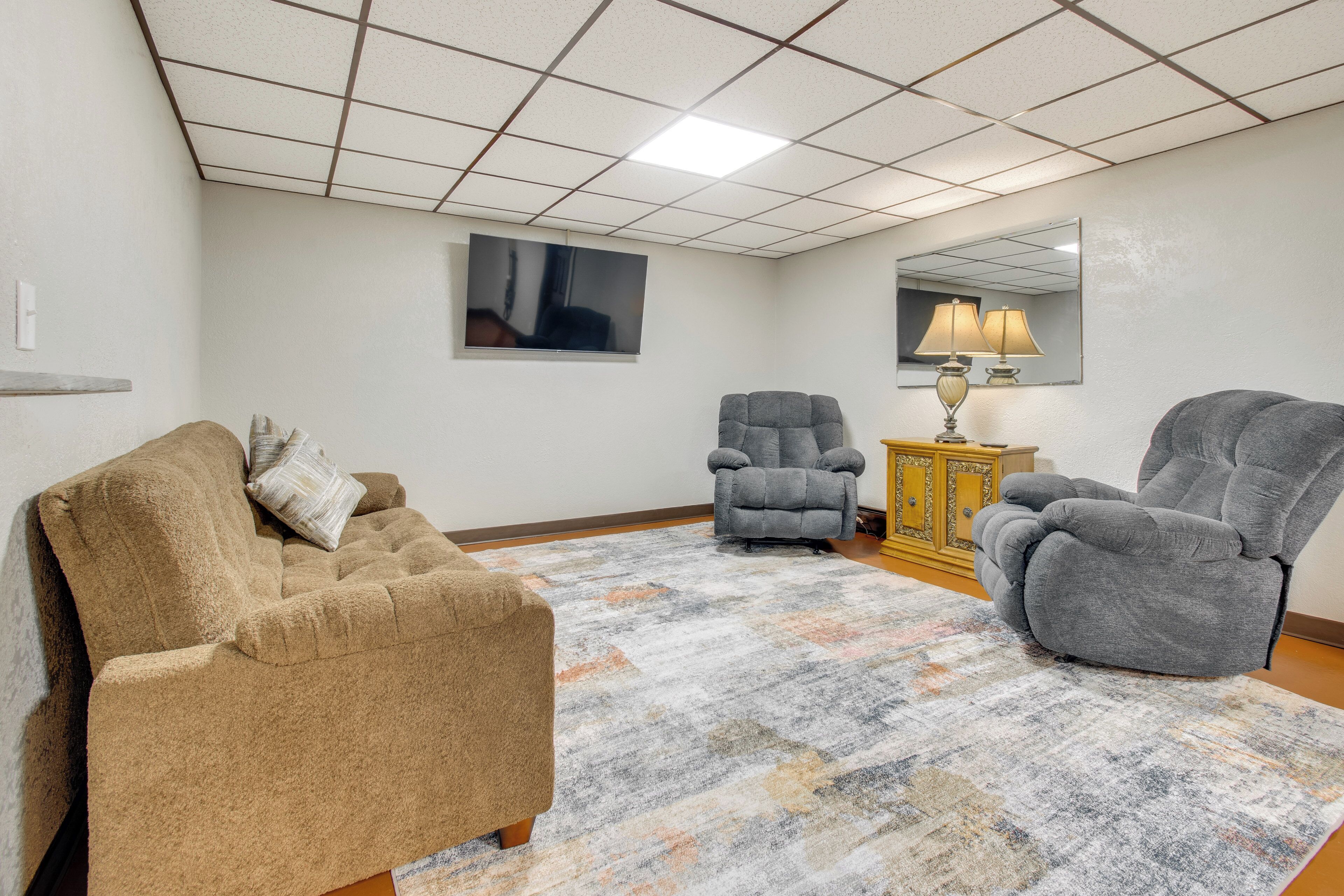 A living room with a beige sofa, two gray armchairs, a patterned rug, a wooden cabinet, and a flat-screen TV mounted on the wall.