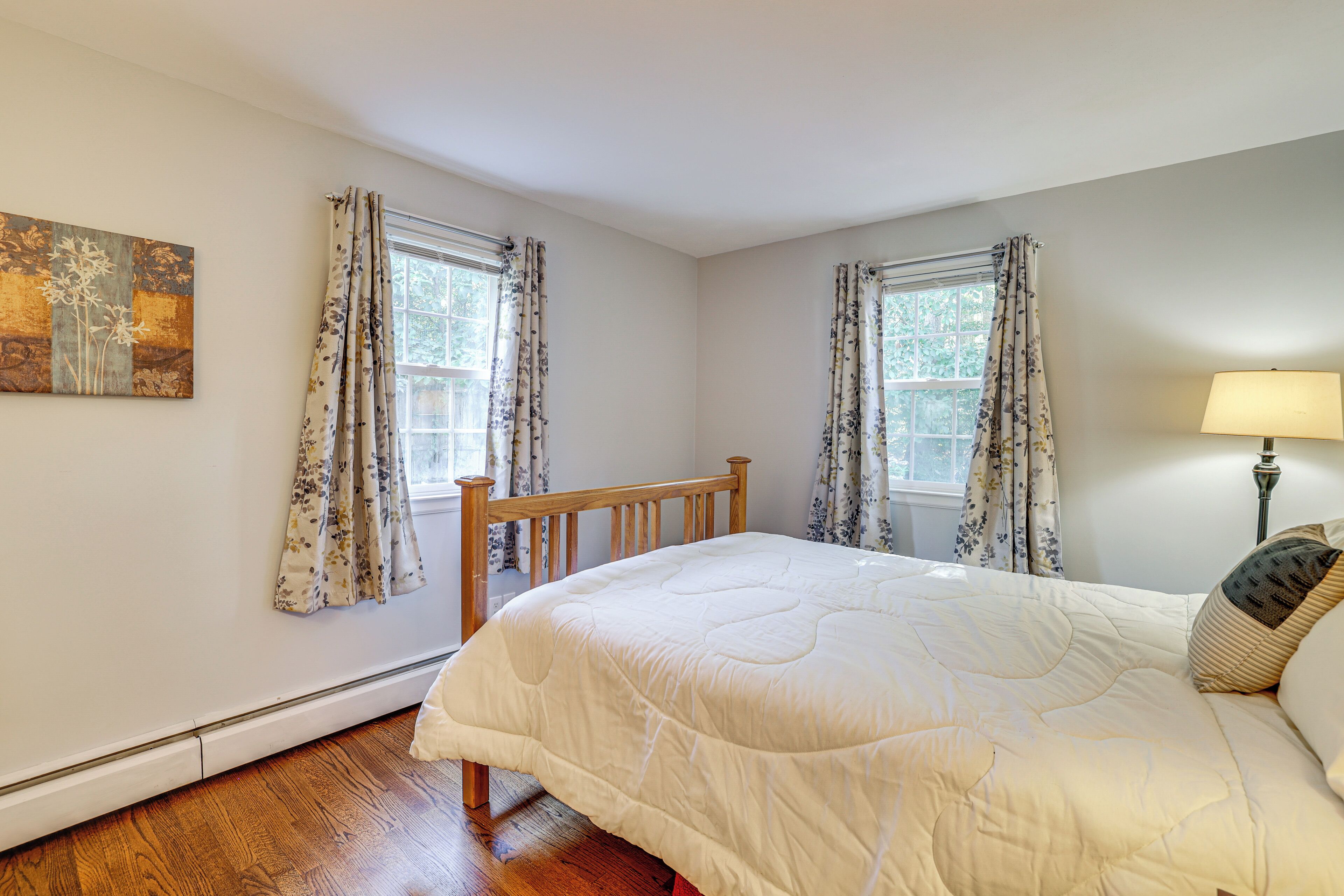 Un dormitorio con una cama de madera, dos ventanas con cortinas florales, una lámpara de noche y un cuadro en la pared.