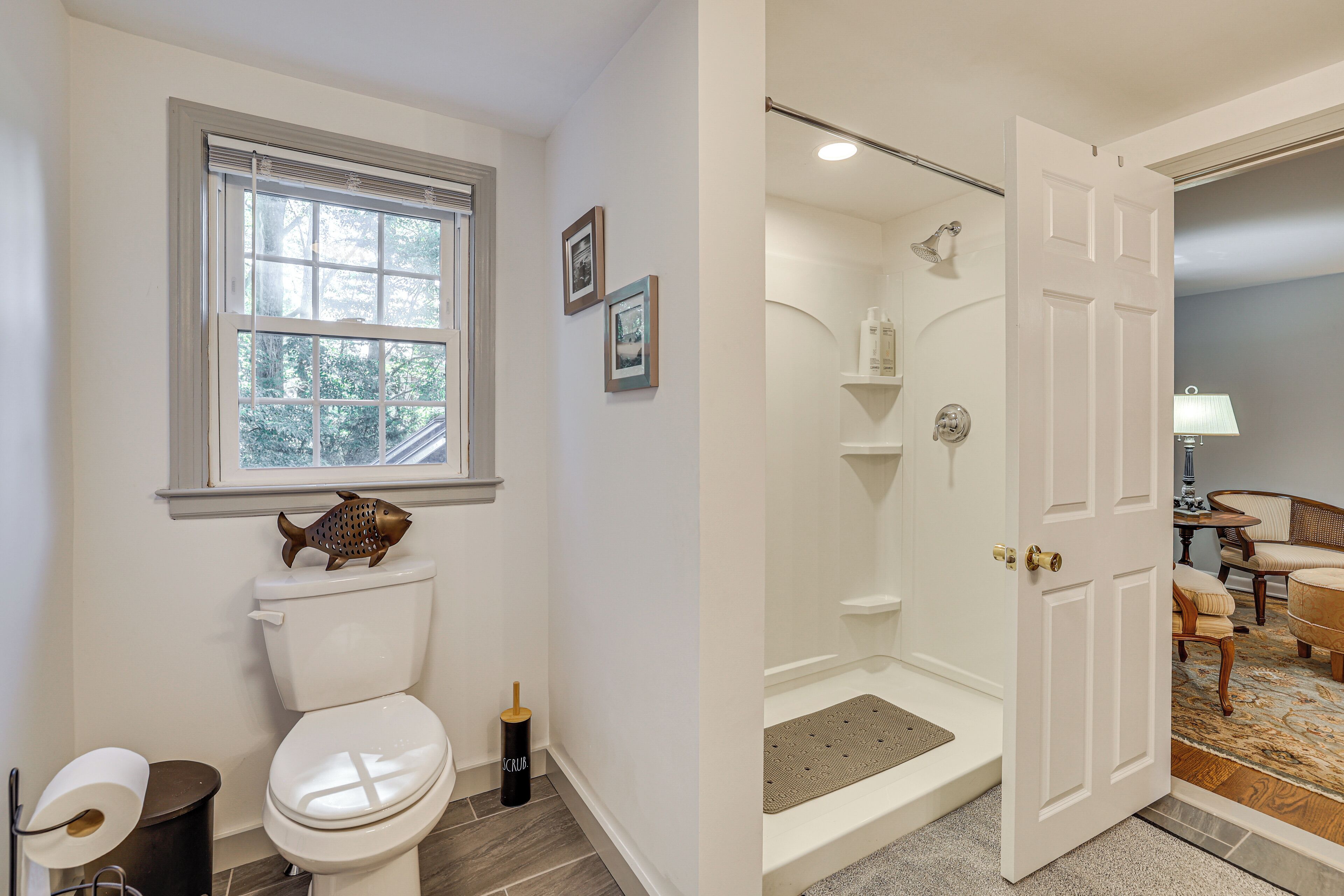 A bathroom with a toilet, a window, a shower, and a door leading to a living area with a chair and a lamp.