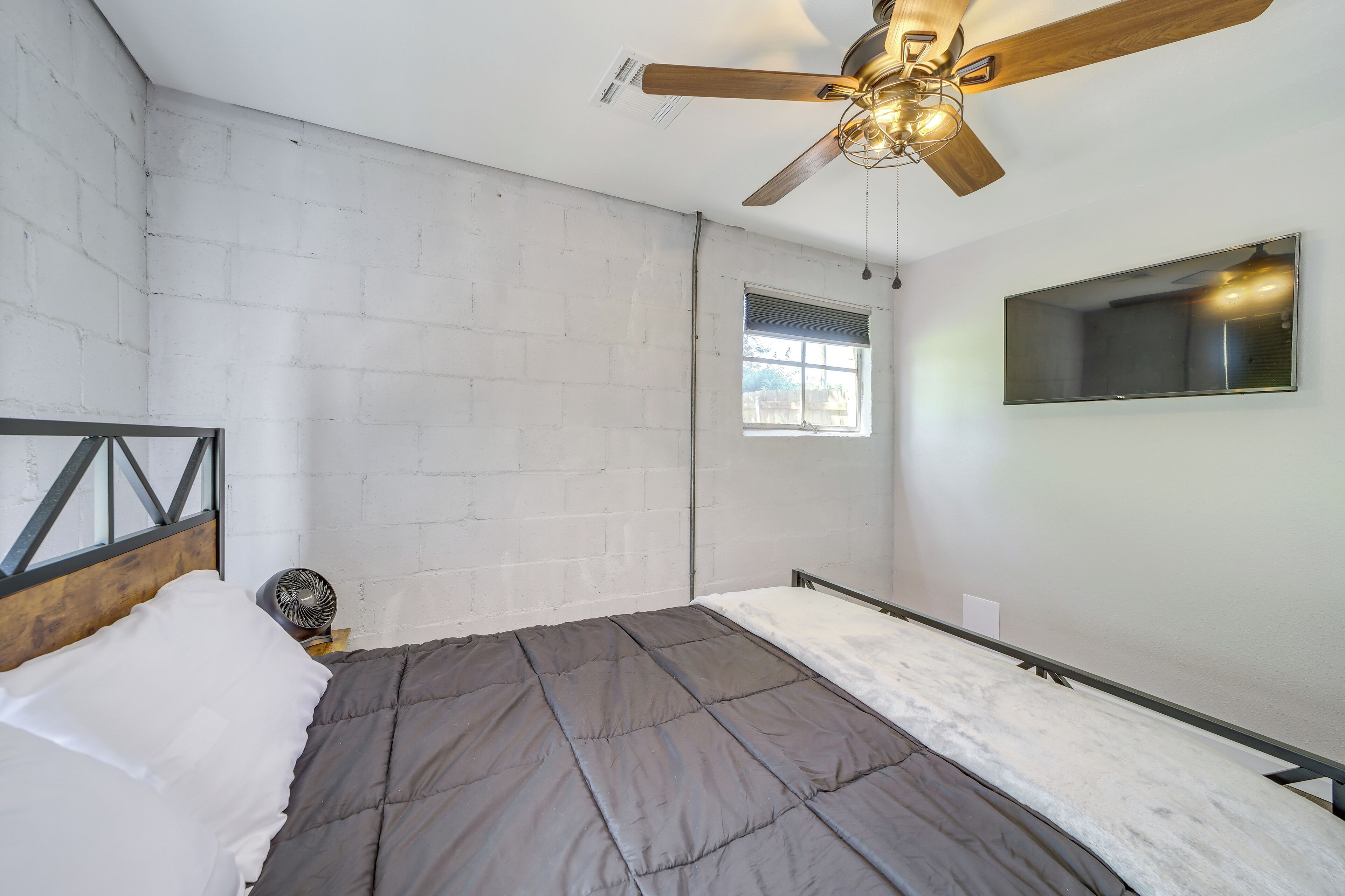 A bedroom with a bed, a ceiling fan, a window, and a TV mounted on the wall.