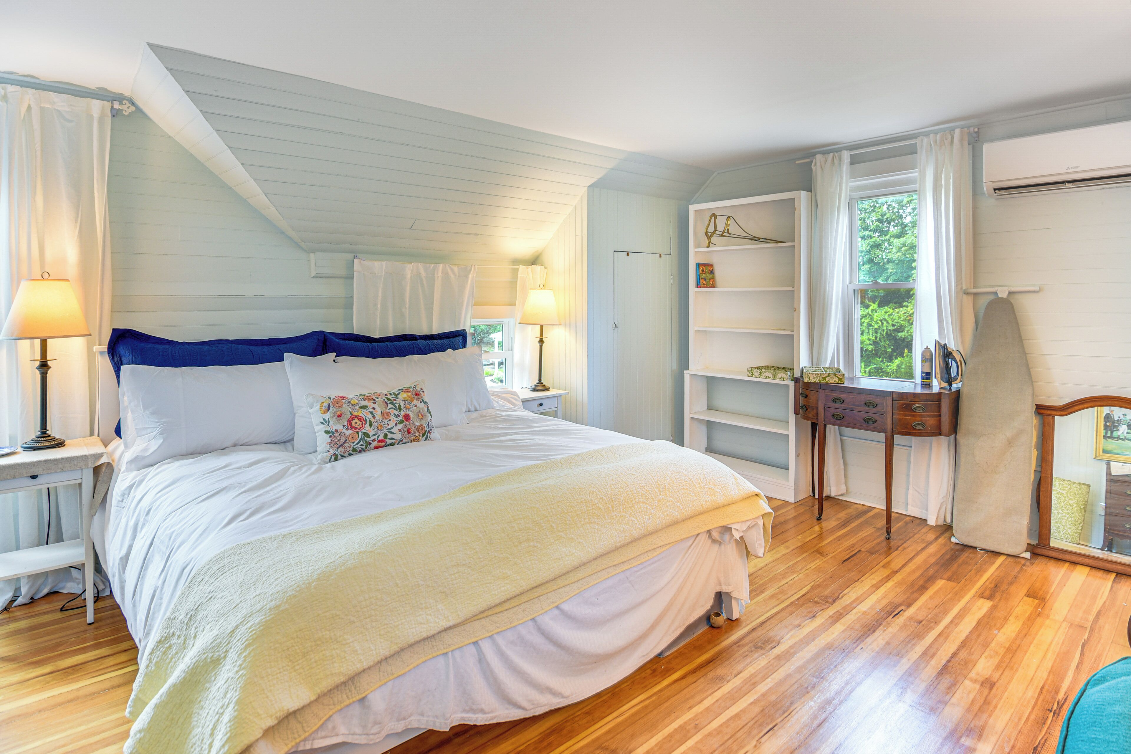 Ein Schlafzimmer mit einem großen Bett, Holzboden, einem Nachttisch mit Lampe, einem Schminktisch und einem Fenster mit Vorhängen.