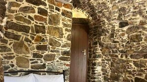 Une pièce aux murs de pierre, avec un lit, une porte en bois et une petite table sur laquelle est posé un vase.