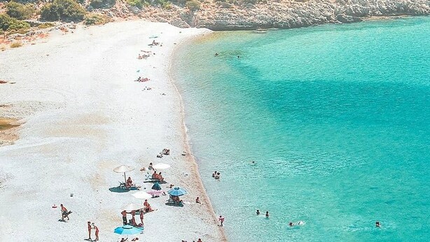 Une vue aérienne d’une plage aux eaux turquoise et limpides, avec un rivage sablonneux et des personnes pratiquant diverses activités.