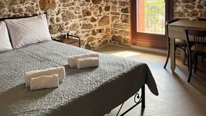 Une chambre avec un mur en pierre, un lit recouvert d’une couette grise, des oreillers blancs et deux serviettes blanches. On y trouve une table à manger avec des chaises et un lustre orné d’abat-jours rouges et jaunes.