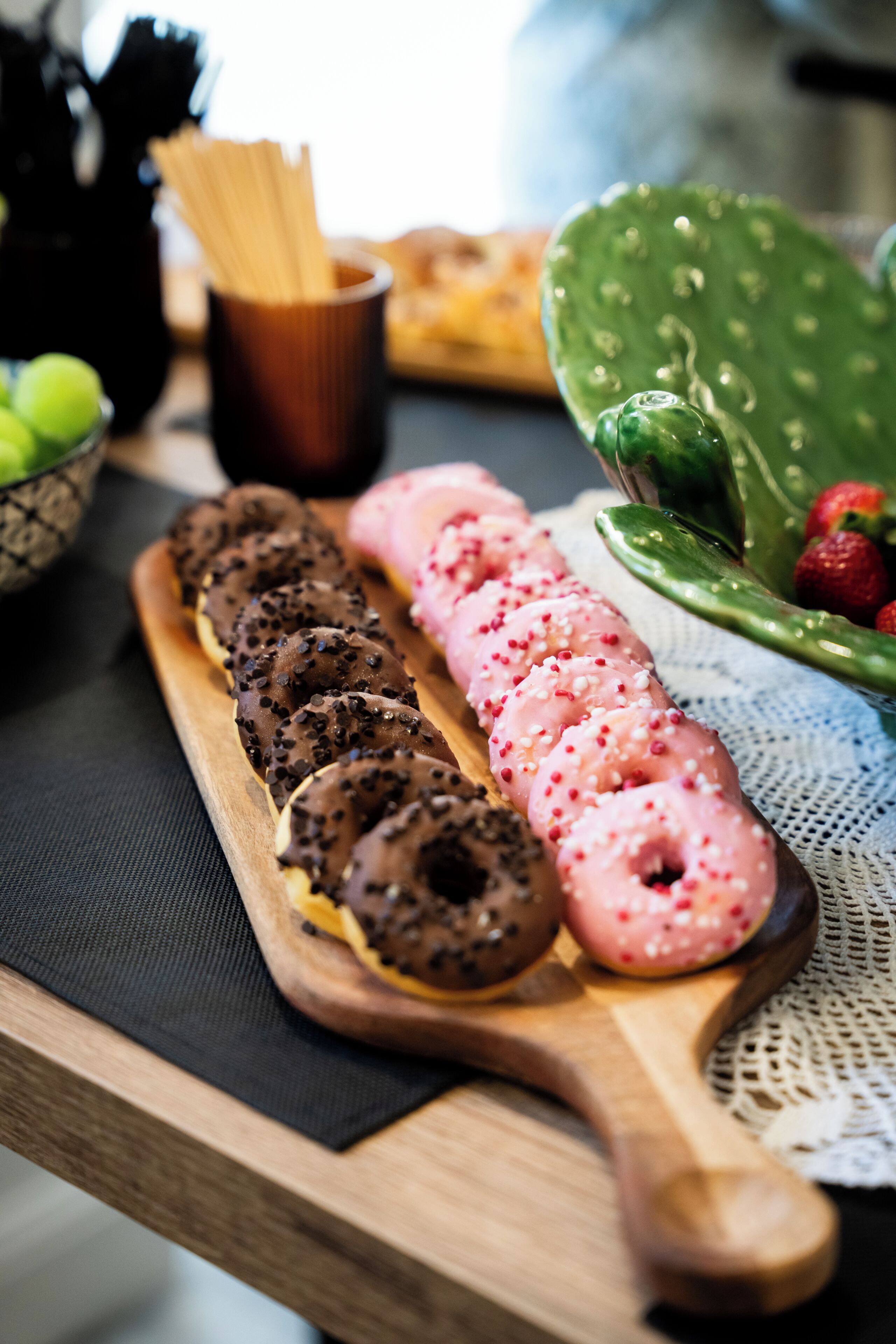 Una tabla de madera con rosquillas de chocolate y rosa, un cactus verde y un bol de fresas.