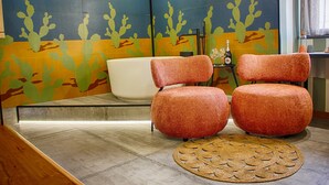 A room with a large bathtub, two orange chairs, a woven rug, and a wall with cactus artwork. - Roby's boutique rooms (Catania)