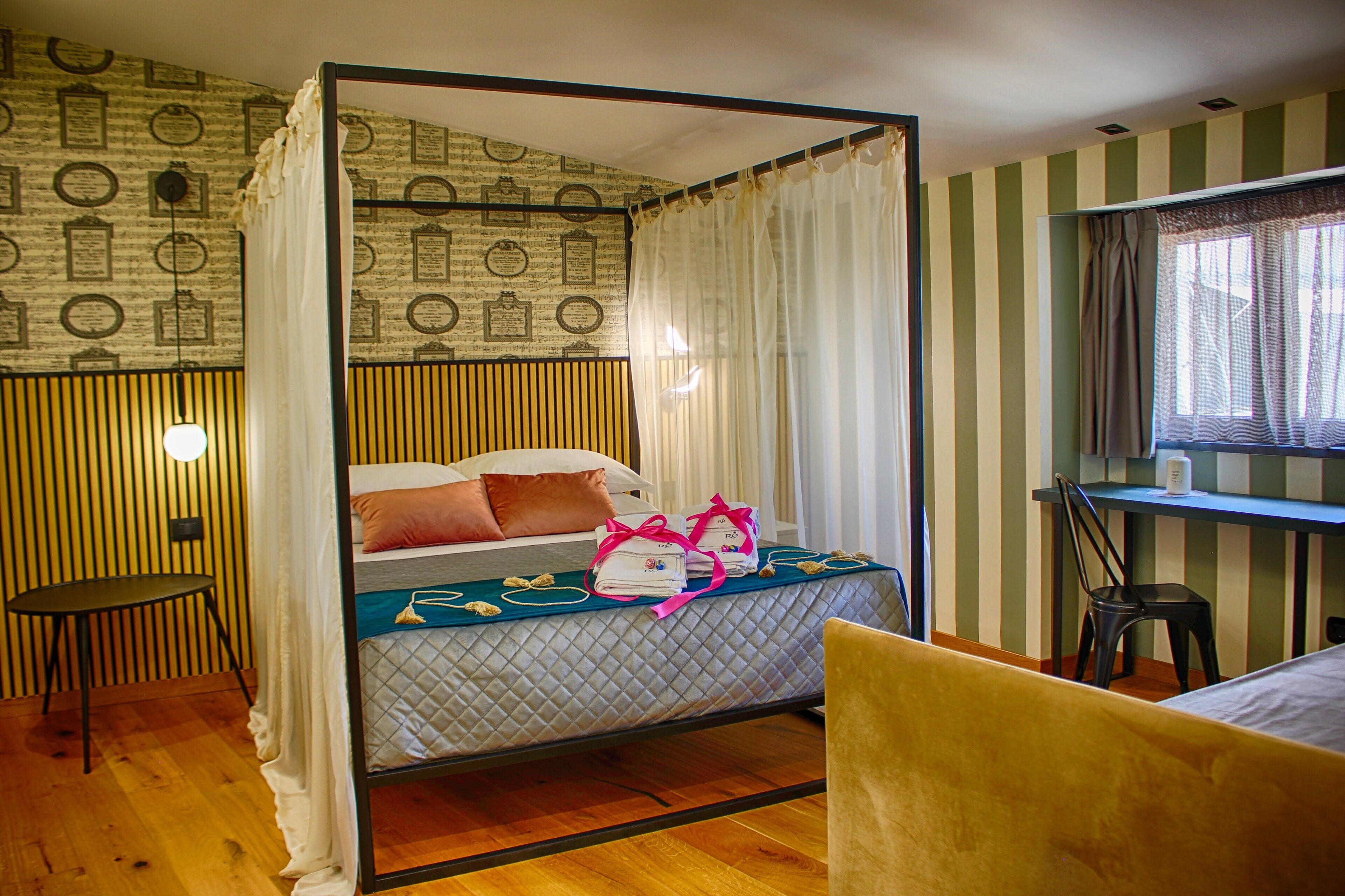 Un lit à baldaquin avec un matelas matelassé, des oreillers et un ruban rose. Une petite table ronde et une chaise se trouvent à gauche du lit. Un bureau avec une chaise est placé à droite du lit. La pièce est décorée de papier peint à rayures et possède une fenêtre avec des rideaux.