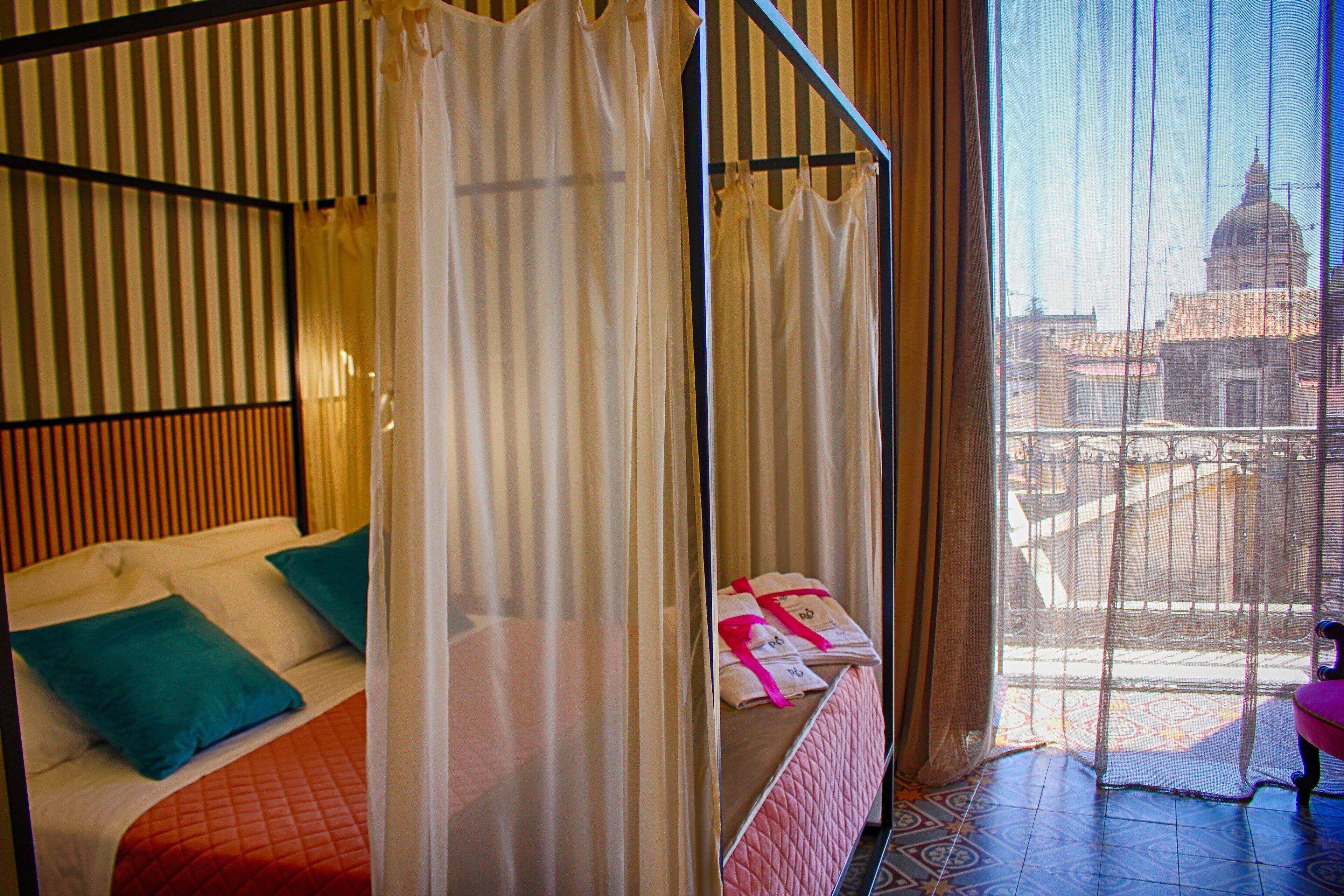 A hotel room with a bed, a window with a view of buildings, and a balcony with a chair.