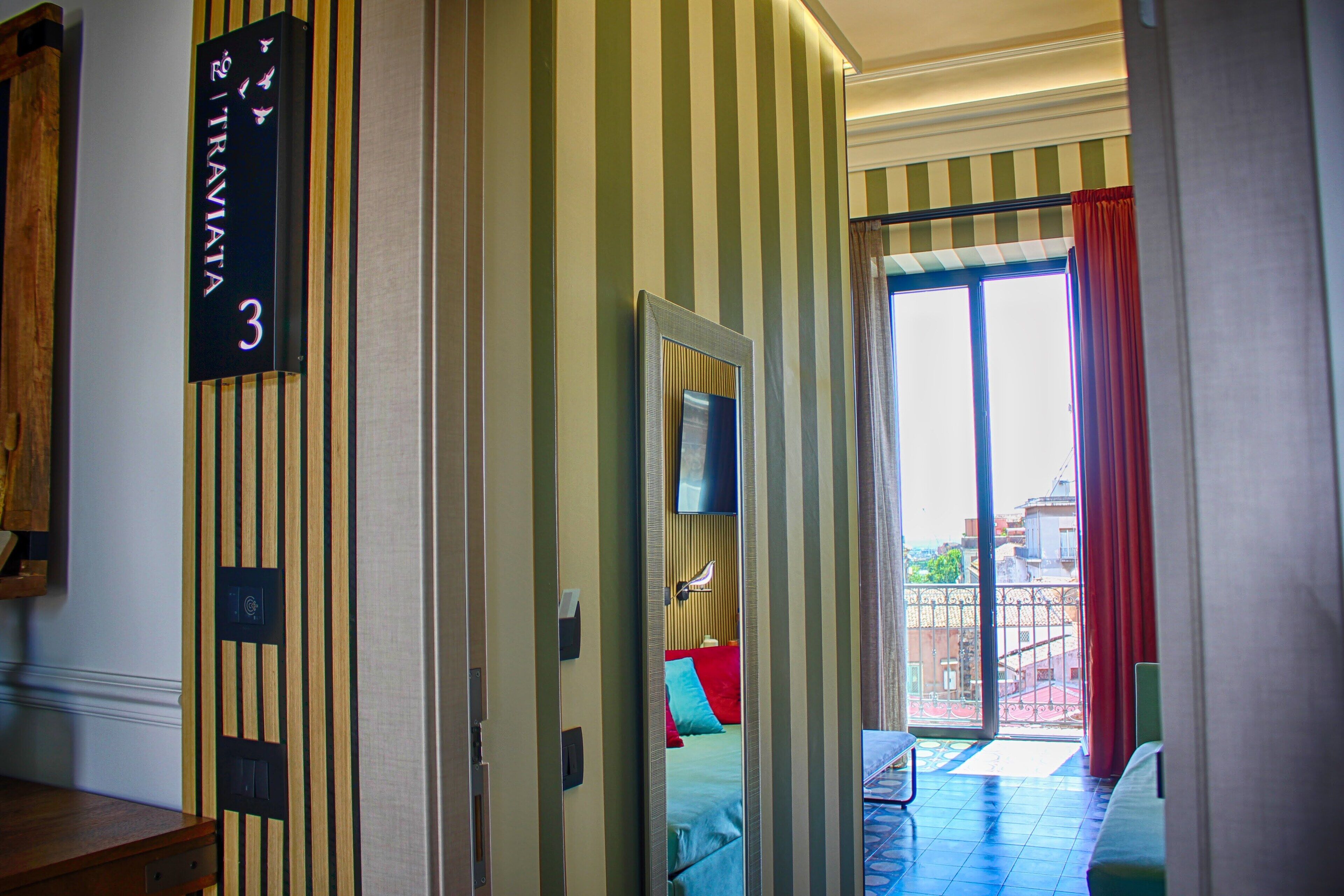 A hotel room with striped wallpaper, a bed with red and blue pillows, a TV on the wall, and a balcony with red curtains.