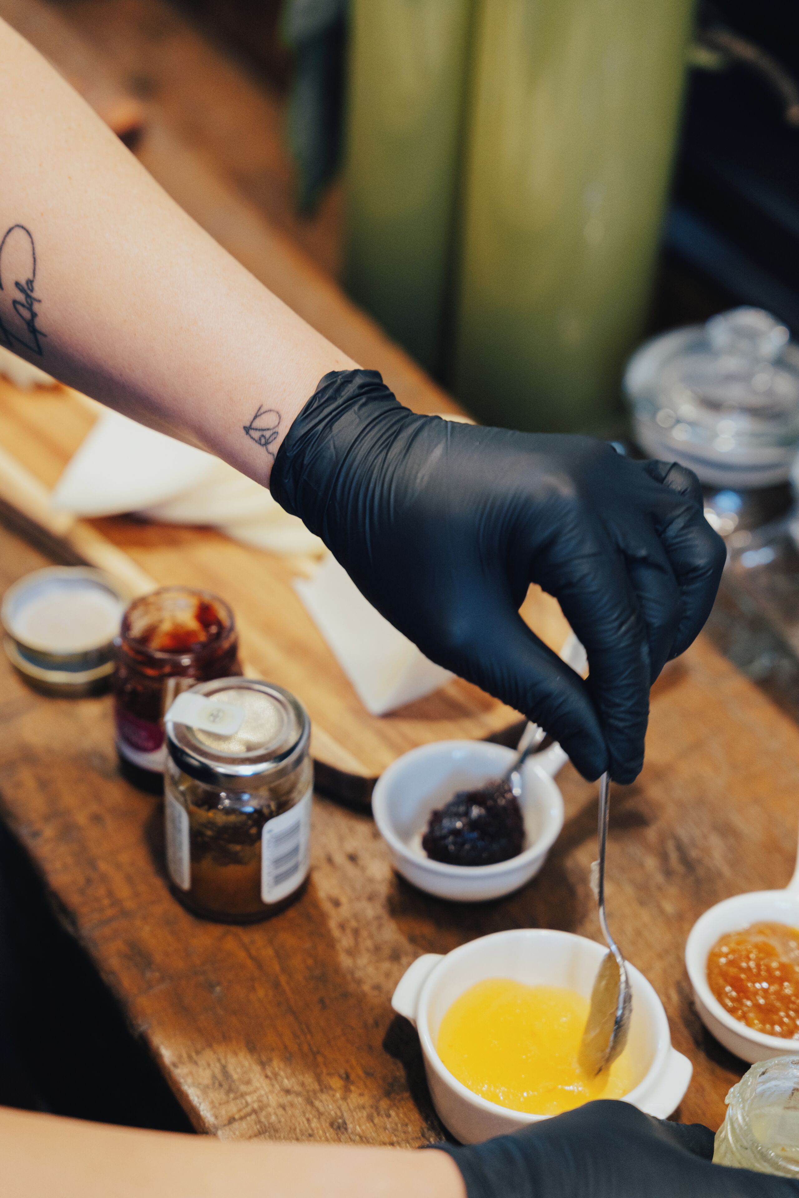 A person wearing black gloves mixing ingredients in a bowl on a wooden table with jars of condiments.