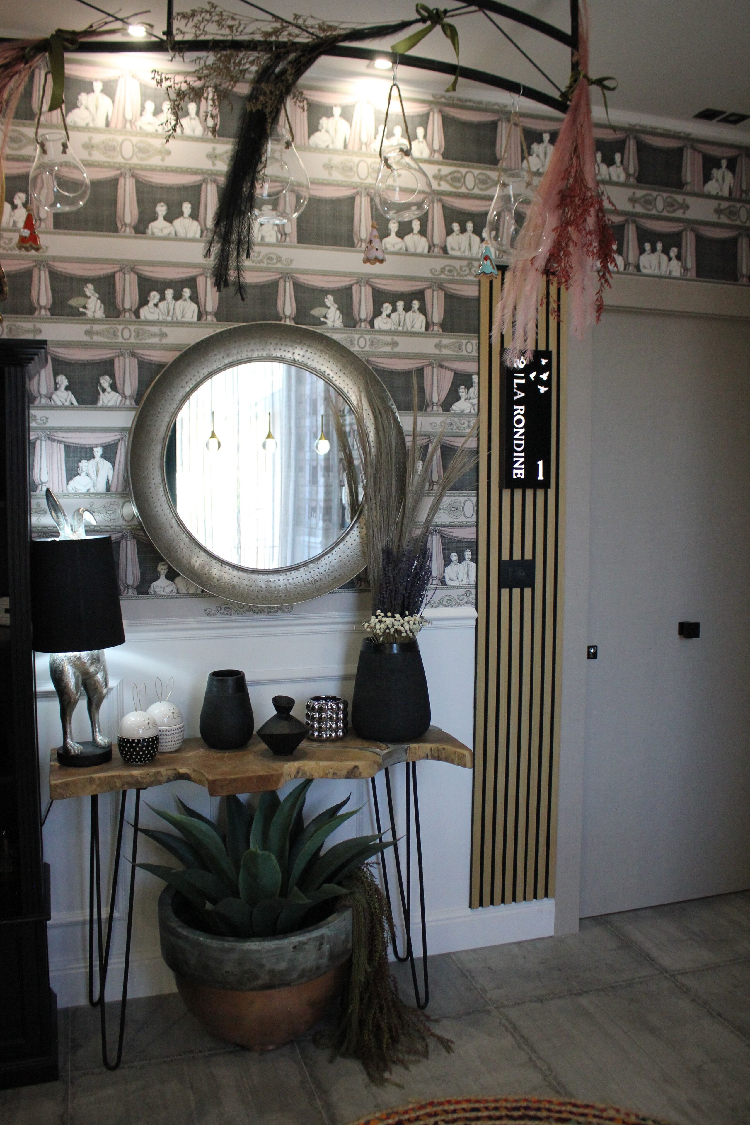 Una habitación con un espejo redondo, una estantería de madera con objetos decorativos, una planta en maceta y un papel tapiz con diseño.