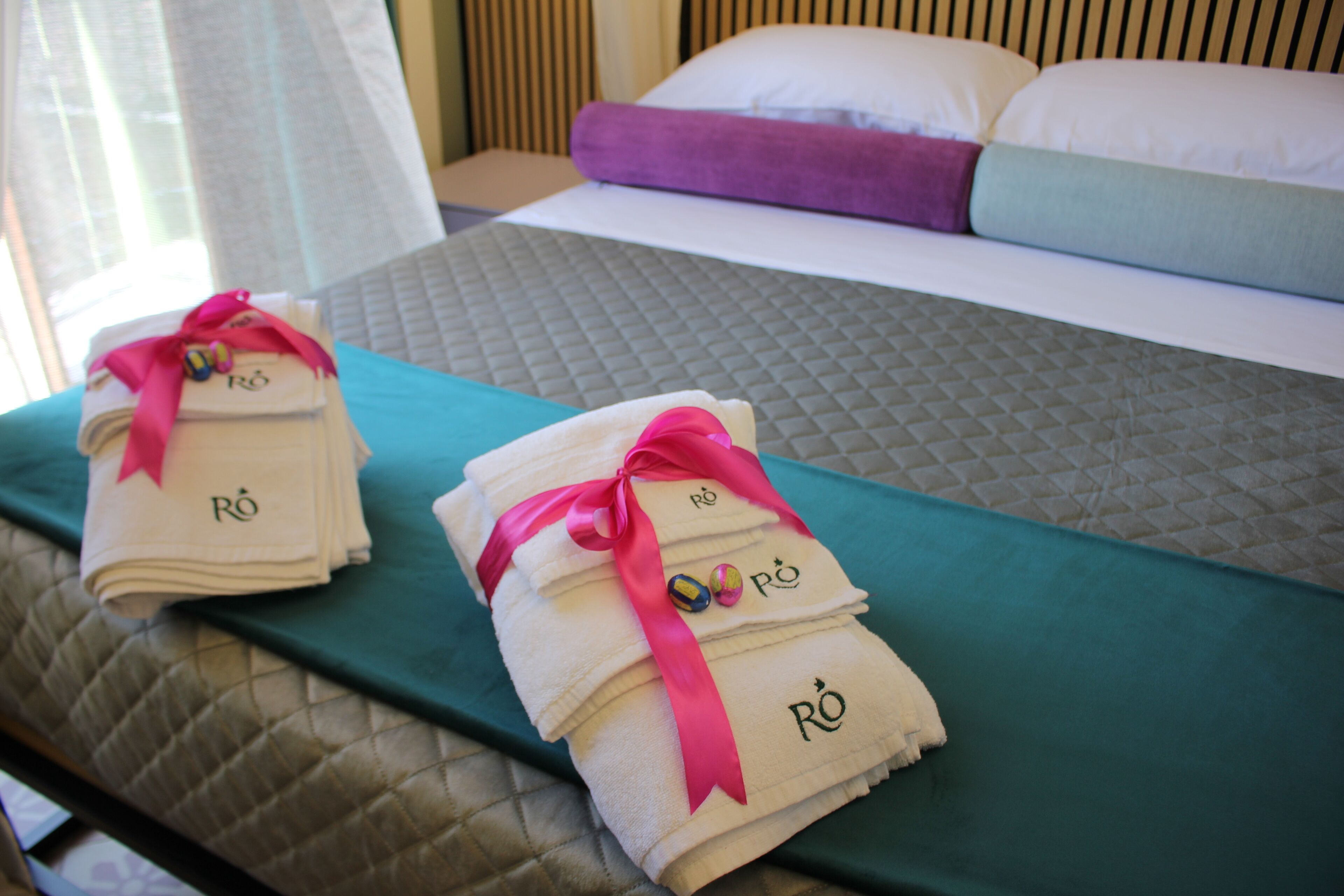 A neatly made bed with a teal bedspread, two folded towels with pink ribbons, and a pillow with a purple cover.