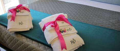 Una cama bien hecha con una colcha color turquesa, dos toallas dobladas con lazos rosas y un almohadón con funda morada.
