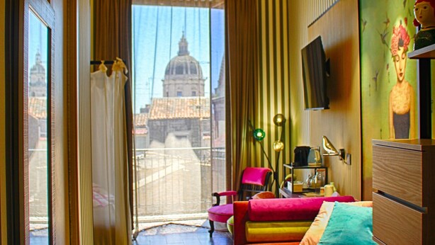 A hotel room with a large bed, a desk, a chair, a TV, and a view of a building with a dome. - Roby's boutique rooms (Catania)