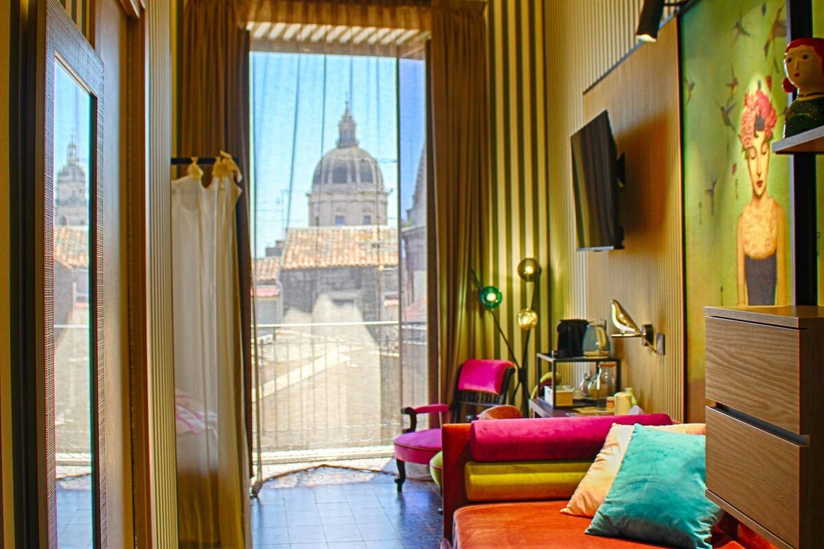 A hotel room with a large bed, a desk, a chair, a TV, and a view of a building with a dome.