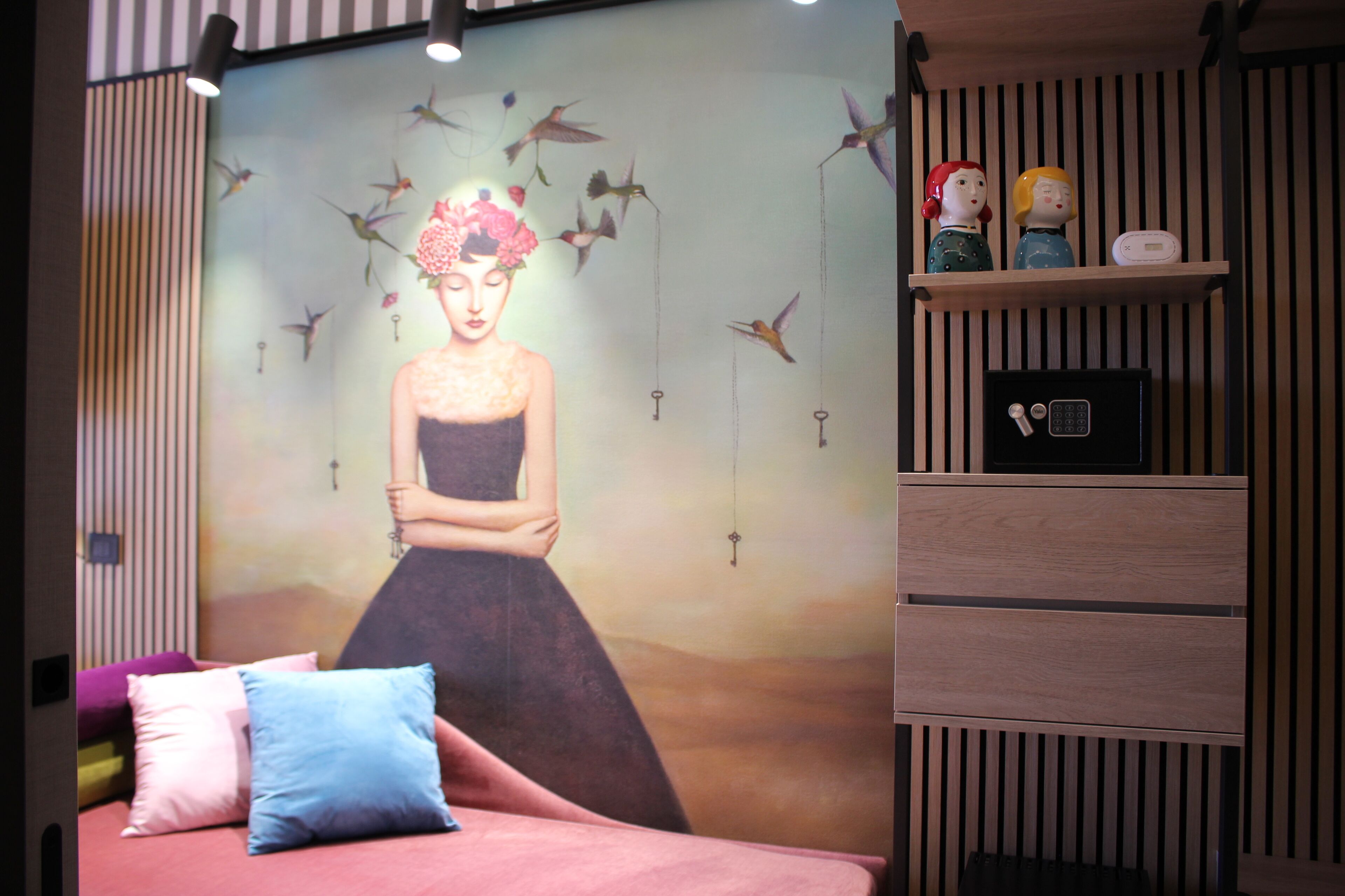 A bedroom with a bed, pillows, a wall mural, a shelf with figurines, and a bedside table.