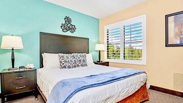 A hotel room with a bed, two bedside tables, a nightstand, a window with blinds, and a wall-mounted artwork.