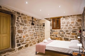 Quarto com paredes de pedra, uma cama, uma mesa de cabeceira com abajur, porta de madeira e uma pequena janela.