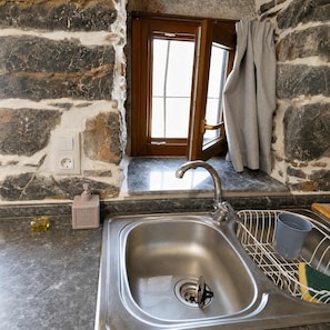 A kitchen with a stone wall, a window with curtains, a sink with a dish rack, and a soap dispenser.