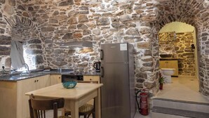 Une cuisine aux murs de pierre, avec un réfrigérateur en acier inoxydable, une table en bois sur laquelle se trouve un saladier, et un évier.