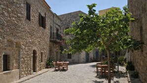 Une ruelle étroite pavée de pierres, bordée de bâtiments en pierre, avec des tables installées à l’extérieur et un arbre qui offre de l’ombre.