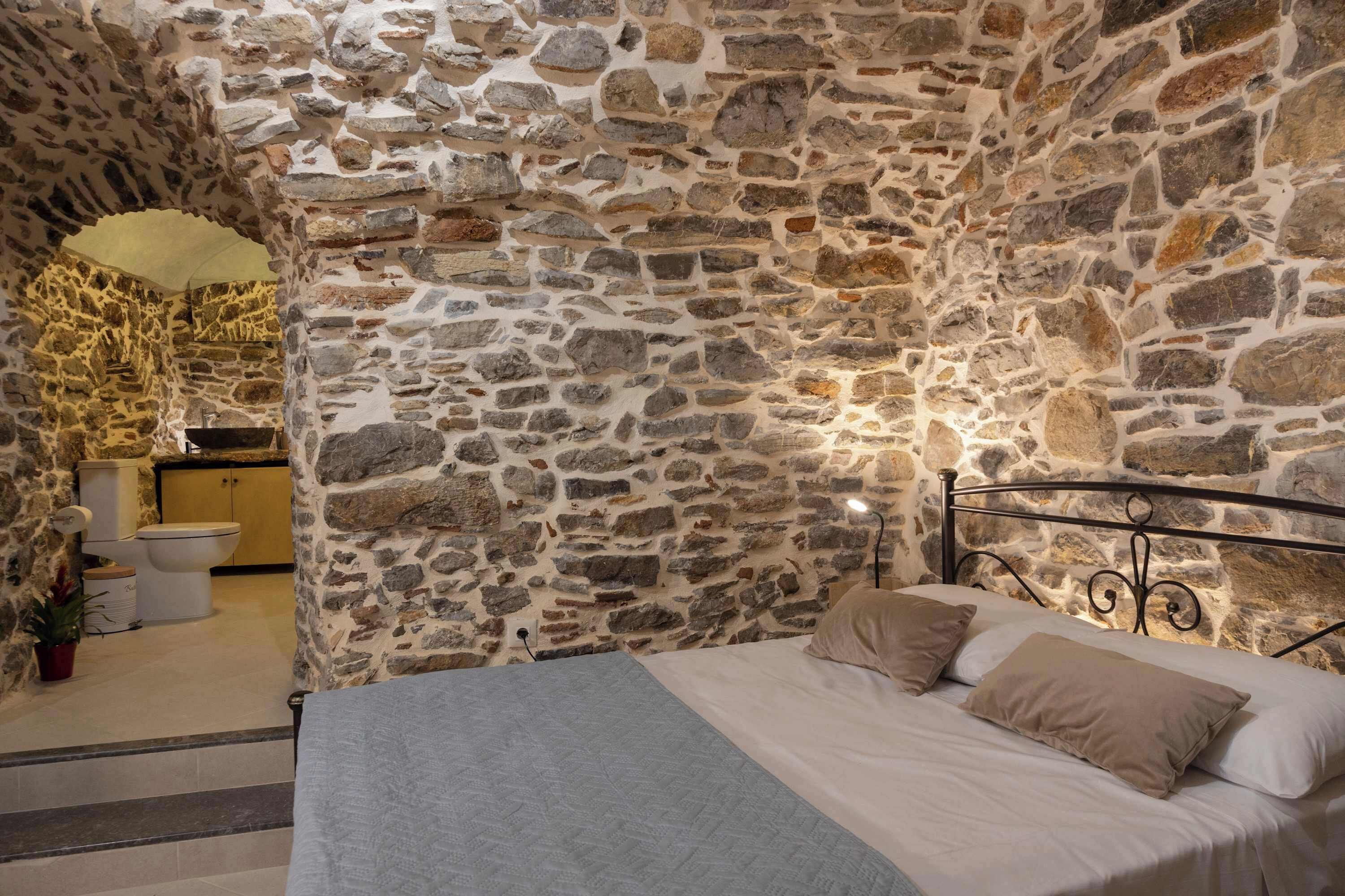 Un dormitorio con pared de piedra, cama con ropa de cama color crema y un baño pequeño con inodoro y lavamanos.