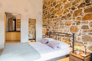 A bedroom with a stone wall, a bed with pillows, a bedside table with a lamp, and a wooden cabinet. - Vlychos Olympi Lodge 2 - Chios Cozy Getaway (Olimpi)