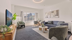 A modern living room with a grey carpet, a blue sofa, a green armchair, and a wooden coffee table. There is a painting on the wall, a plant on the desk, and a window with curtains. - 1 Bedroom Apartment with City Skyline Views (Pyrmont)