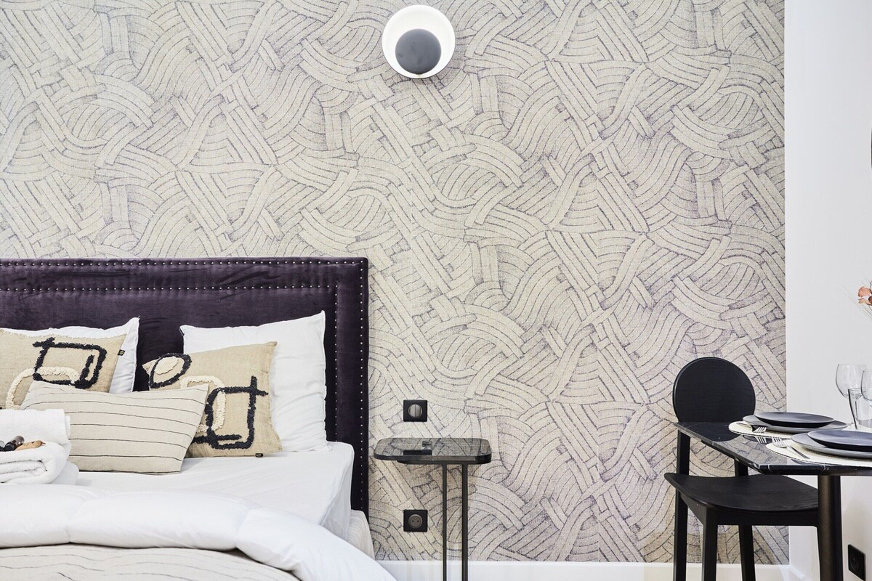 Un lit bien fait, avec une tête de lit sombre, une petite table de chevet et un mur recouvert d’un papier peint à motifs géométriques.