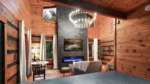Un interior de cabaña acogedor con chimenea, paredes de madera, zona de comedor y un amplio ventanal.