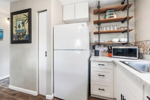 Eine kompakte Küche mit einem weißen Kühlschrank, einer Mikrowelle und einem Spülbecken. Darüber befinden sich Holzregale mit Gläsern und Küchenutensilien.